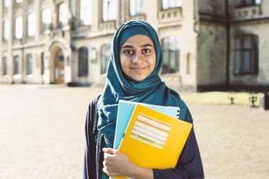 Üniversitenin yanında kitap tutan gülümseyen müslüman genç kız öğrenci. Türbanlı mutlu Arap kızı. Eğitim asyalı kadın.  