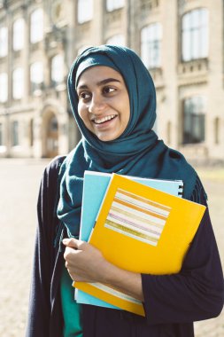 Üniversitenin yanında kitap tutan gülümseyen müslüman genç kız öğrenci. Türbanlı mutlu Arap kızı. Eğitim asyalı kadın.  