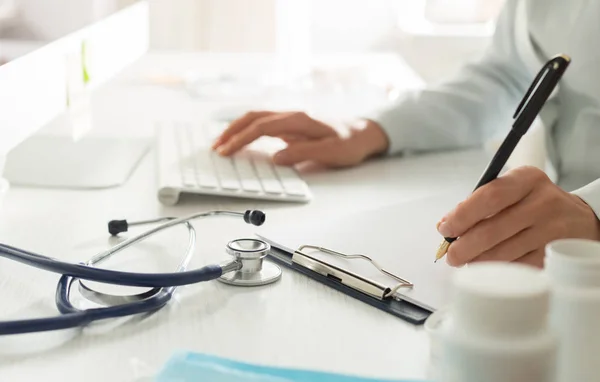 A female doctor writes something in a notebook and works at a computer while sitting at a work desk.