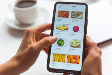 Akıllı telefon kullanarak evde oturan bir kadın internet mağazasından yiyecek satın alıyor. Cep telefonu ekranındaki kadın eli malları uzaktan seçer.