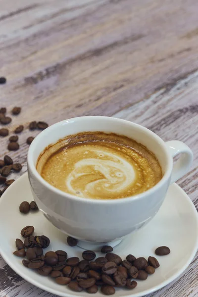 Taza blanca con café negro con leche. Unos granos de café en una mesa de madera. vista vertical de la bebida con café. Copiar espacio de cerca . — Foto de Stock
