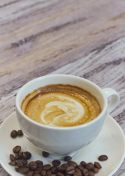 Kubek biały z czarną kawę z mlekiem. Garść ziaren kawy na drewnianym stole. Widok pionowy napoju z kawy. Miejsce z bliska. — Zdjęcie stockowe