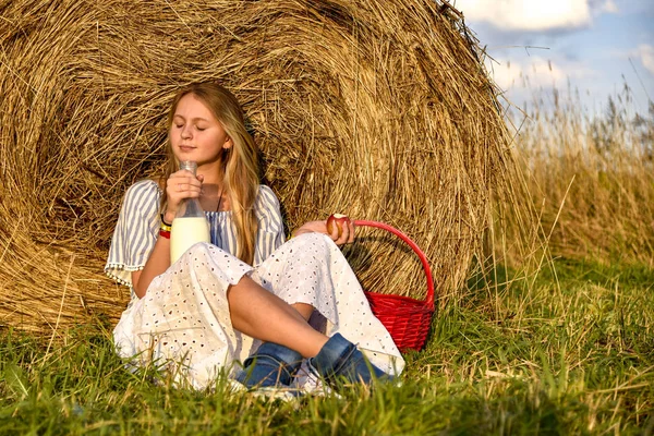 Een meisje met lang haar zit op de grond in een veld in de buurt van een hooiberg en drinkt melk uit een grote fles — Stockfoto