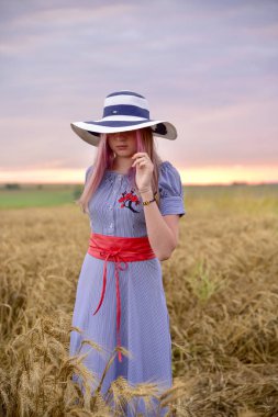 Bir kız mavi bir elbise ve hafif bir şapka bir buğday alan sarı ortasında duruyor
