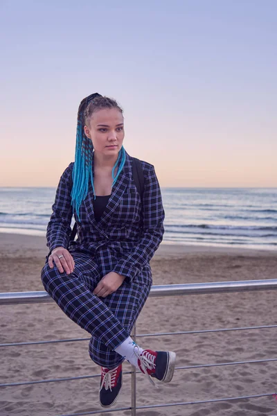 青いドレッドロックとチェック柄のスーツを着た少女は ビーチと海に対する鉄の手すりに座っている — ストック写真