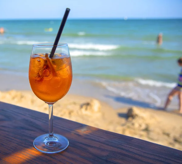 A glass with an orange cocktail an aperol with a black straw stands on a bar counter against the background of the sea and holidaymakers.