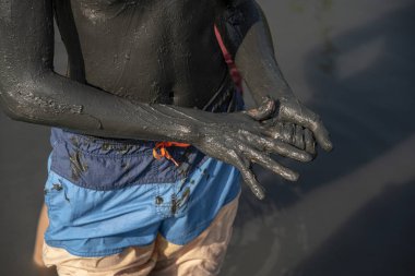 Bir gencin vücudu ve kolları, kalın siyah şifa çamur ile bulaşmış, üzerinde banyo şort var. Şifalı çamuru olan bir arka plan gölü gibi..