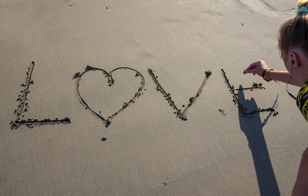 The girl writes the word love on the sea sand, the letter o is drawn in the form of a heart.