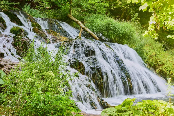 Şelalenin fotoğrafı, uzun bir deklanşör hızında çekilmiş. Bulanık su akışı, güneş ışığı, çevrede yeşillik çok.