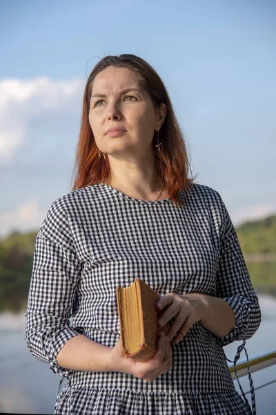 Жінка, одягнена в плетену туніку і несе книгу, стоїть навпроти річки, дивлячись на відстань . — стокове фото