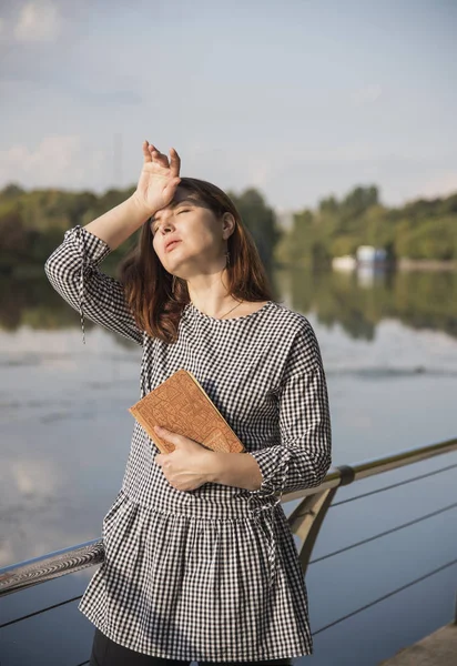 Сорокалетняя женщина в клетчатой тунике стоит с книгой в руках у реки — стоковое фото