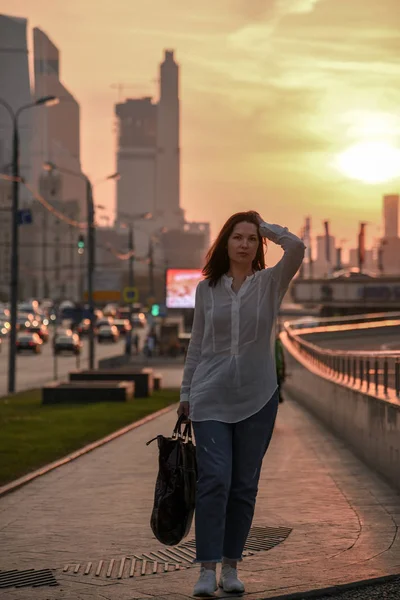 Uma menina em uma blusa branca e carregando um saco grande está andando pela cidade contra o céu do por do sol . — Fotografia de Stock