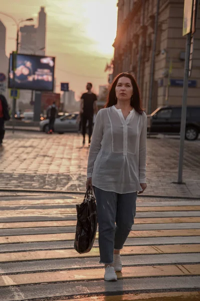 A woman in a white blouse and carrying a large bag walks through the city in the haze of sunset.