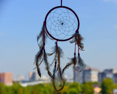 A Dreamcatcher suspended from a window.Outside, the sky is overcast and the city is blurred. clipart