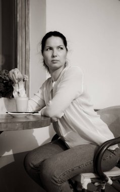 Bir kadın açık bir kafede bir masada oturur ve bir kaşıkla kahve karıştırır. Siyah beyaz fotoğraf.