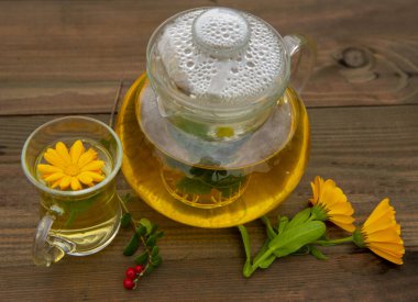 Şeffaf çaydanlık ve ahşap arka planda bir fincan bitki çayı. Calendula çiçeklerinin yanında