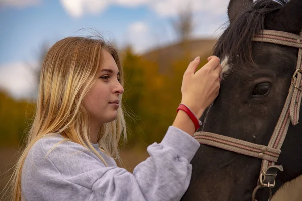 En ung flicka med blont hår smeker försiktigt den bruna hästens munkorg. — Stockfoto