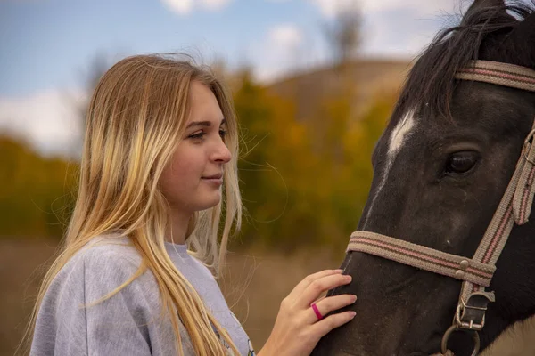 En ung flicka med blont hår smeker försiktigt den bruna hästens munkorg. — Stockfoto