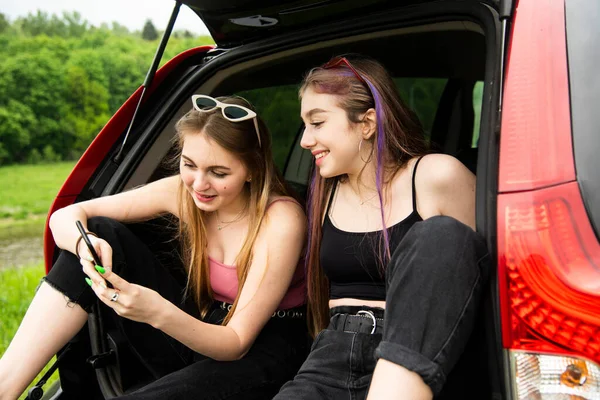两个留长发的女性朋友 一个金发碧眼 一个黑发 坐在汽车后备箱里 看着电话 — 图库照片