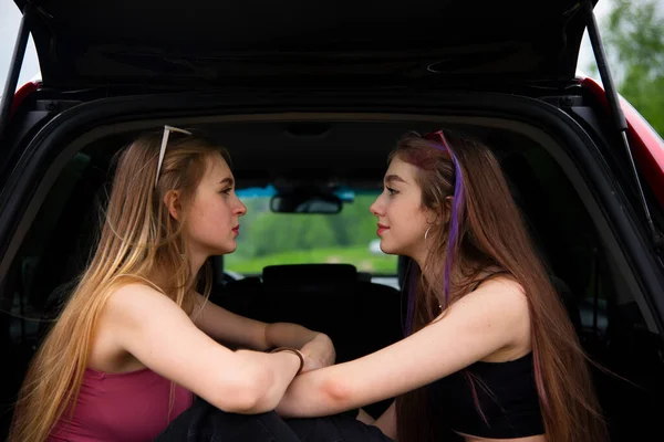 Dos hermosas chicas se sientan en el maletero de un coche enfrente de la otra  . — Foto de Stock