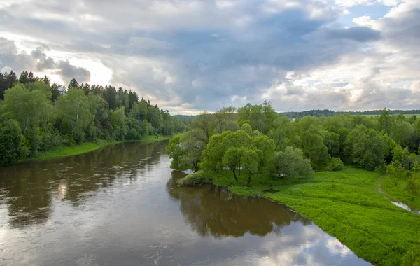 Summer landscape. A dark river flows among green banks under a dark, cloudy sky.