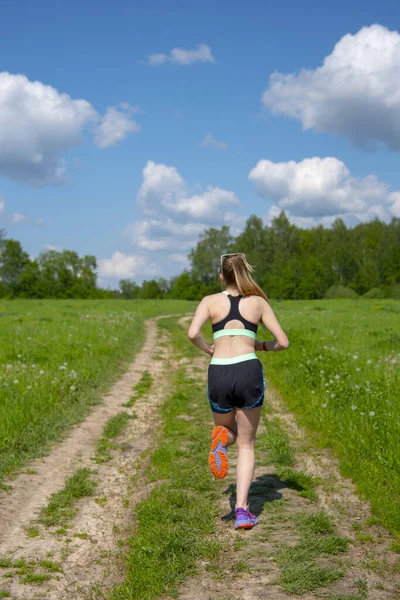 The girl is engaged in running. She runs along a road that runs through fields under a blue cloudy sky on a summer day.