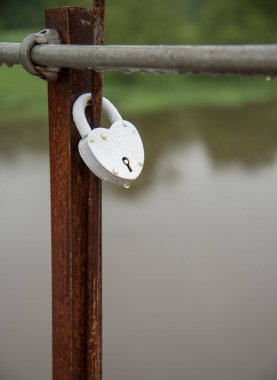 Dikey fotoğraf. Kalp şeklindeki kilit köprüye sabitlenmiş. Doğanın arka planına karşı. Metal kilidin üzerinde yağmur damlaları var..
