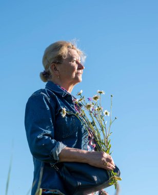 70 yaşında, güneş ışığıyla aydınlanan yaşlı bir kadın bir buket papatyayla mavi gökyüzüne karşı poz veriyor..