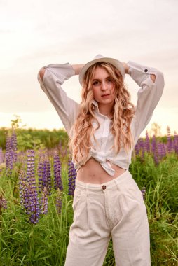 Beyaz elbiseli ve şapkalı bir kız, uzun sarı saçlı, çiçek açan lupinlerin arkasında poz veriyor..