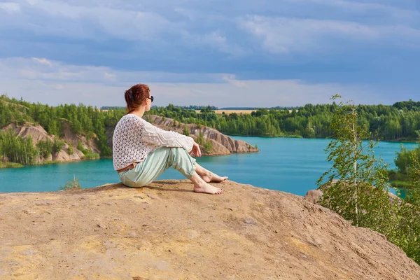 Uma mulher com cabelo vermelho e óculos escuros senta-se em uma montanha alta acima de um lago azul brilhante  . — Fotografia de Stock