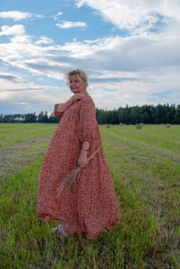 Uzun kır elbiseli, sarı saçlı yaşlı bir kadın samanlıkta poz veriyor..