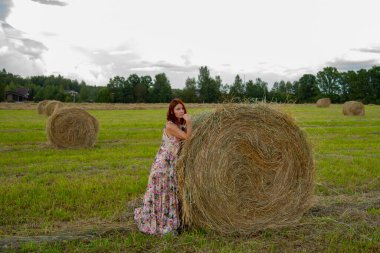 Uzun, zarif bir yaz elbisesi içinde kızıl saçlı orta yaşlı bir kadın samanlığa yaslanıyor..