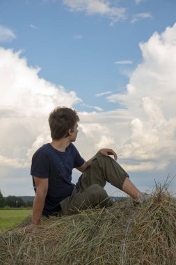 Yakışıklı bir genç adam mavi bulutlu bir gökyüzüne karşı samanlıkta oturuyor..