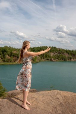 Uzun sarı saçlı, açık renk elbiseli ve çıplak ayaklı bir kız nehrin yukarısındaki bir uçurumda elleri havada duruyor..