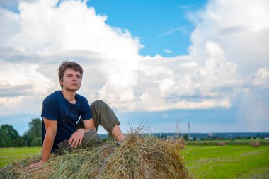 Yakışıklı bir genç adam mavi bulutlu bir gökyüzüne karşı samanlıkta oturuyor..