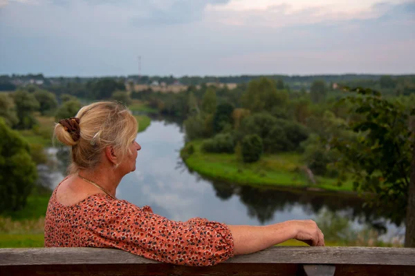 An elderly 65-year-old woman sits alone on a bench on a high Bank above the river with her back to the camera.