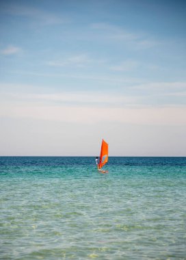 Mavi deniz ve gökyüzüne karşı kızıl yelkenli altında bir sörfçü.