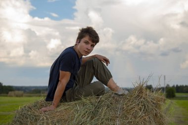 Yakışıklı bir genç adam mavi bulutlu bir gökyüzüne karşı samanlıkta oturuyor..