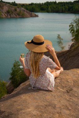 Hasır şapkalı ve yazlık elbiseli sarışın bir kız gölün yukarısındaki yüksek bir tepede bir bardak şampanyayla oturuyor..