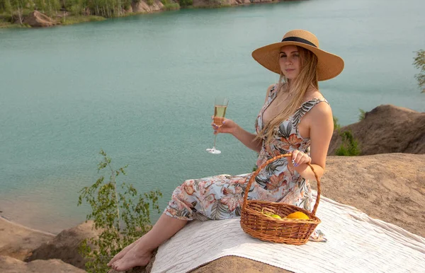 A girl with long blond hair in a hat and a smart sundress sits with a glass of champagne on a hill with her back to the camera.There is a fruit basket next to it.