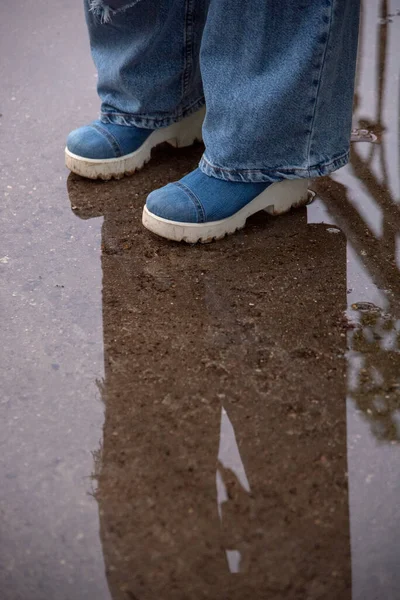 Women\'s feet in denim boots on a high wedge on the background of wet asphalt.