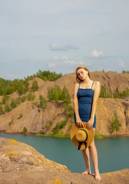 Sarı saçlı, kot elbise giyen genç bir kız mavi gölün üzerindeki yüksek bir tepede duruyor..