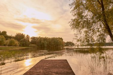 Sonbahar akşamı manzarası. Güneş, ağaçlarla çevrili olarak gölün yüzeyine yansıyor. Ön planda tahta köprüler var..