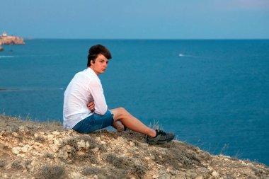 Beyaz gömlekli ve kot şortlu genç bir adam denizin üstündeki bir kayanın üzerinde oturuyor..