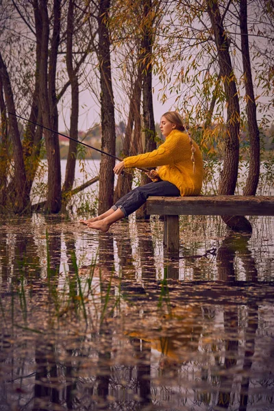 Een jong meisje in een gele trui zit op de brug over het meer en vangt vis. — Stockfoto
