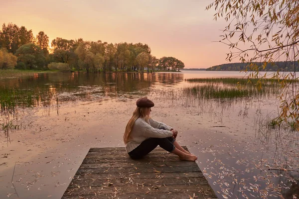 Een blootsvoets meisje met lang blond haar en een pet zit op de brug bij het meer tegen de achtergrond van zonsondergang . — Stockfoto