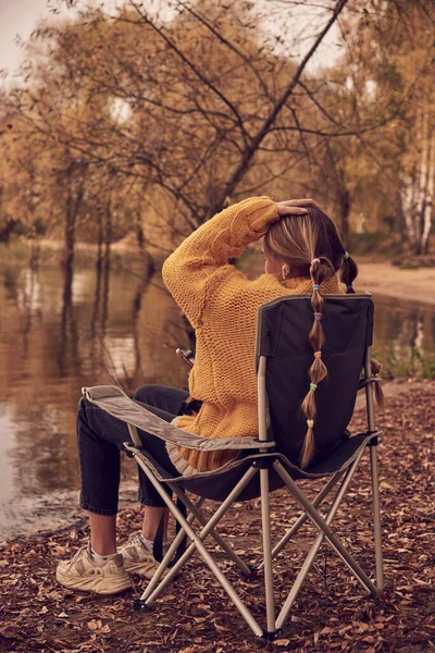 Een tienermeisje in een felgele trui zit in een kampstoel aan de oever van een meer.. — Stockfoto