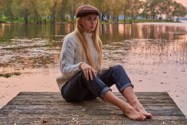 Uzun sarı saçlı ve şapkalı çıplak ayaklı bir kız gölün kenarındaki köprüde gün batımının arka planında oturuyor. .