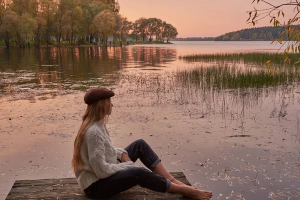 Uma menina descalça com cabelo loiro longo e um boné se senta na ponte junto ao lago contra o fundo do pôr do sol . — Fotografia de Stock
