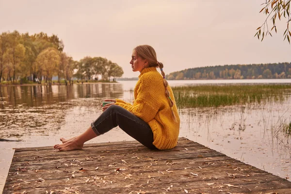 Uma menina descalça com o cabelo em tranças se senta em uma ponte de madeira junto ao lago contra o pôr do sol. A menina está segurando uma xícara com uma bebida. — Fotografia de Stock